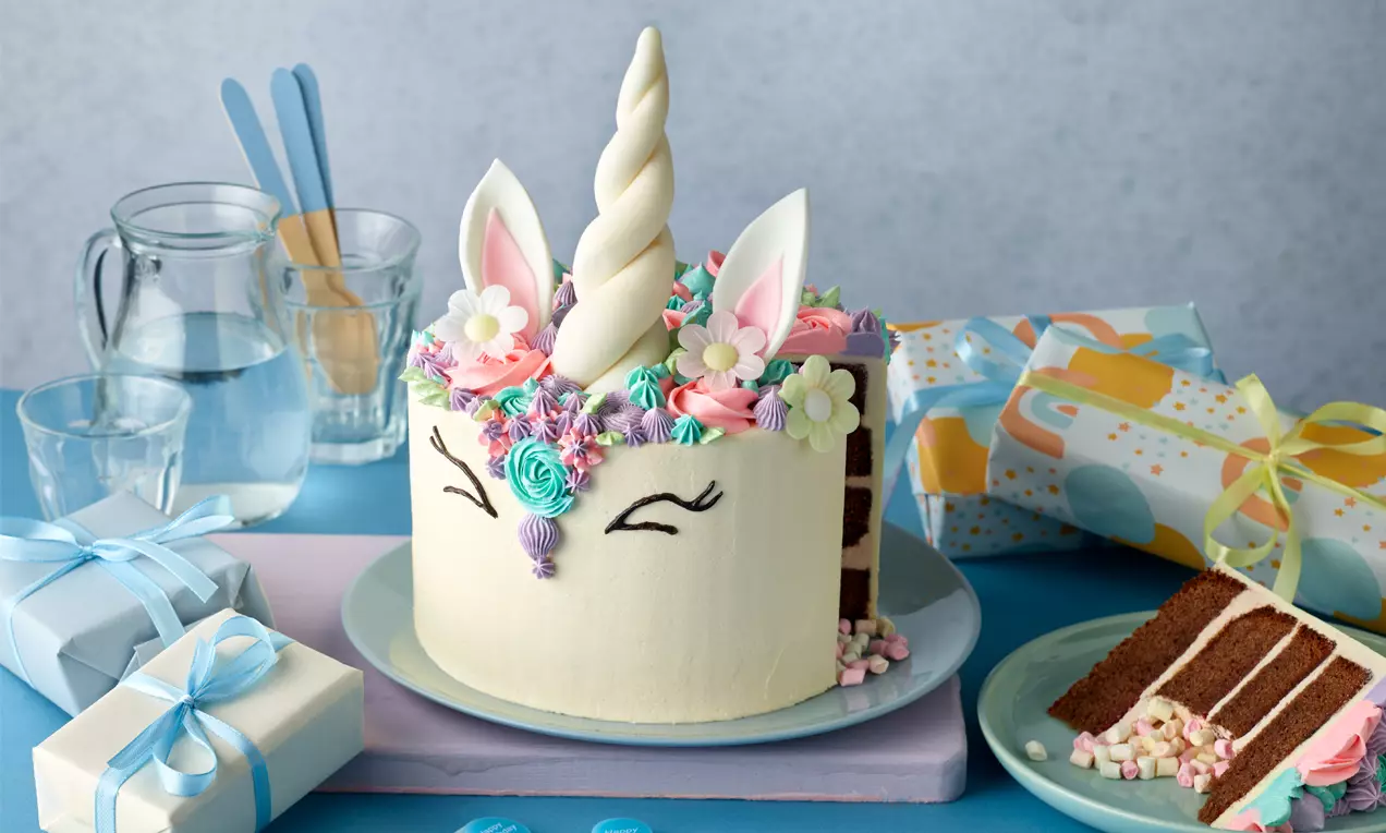 Runde Einhorn-Torte mit cremeweißer Oberfläche, bunten Zuckergussblumen, aufgesetztem Horn, Ohren und aufgemaltem Gesicht auf einem Teller