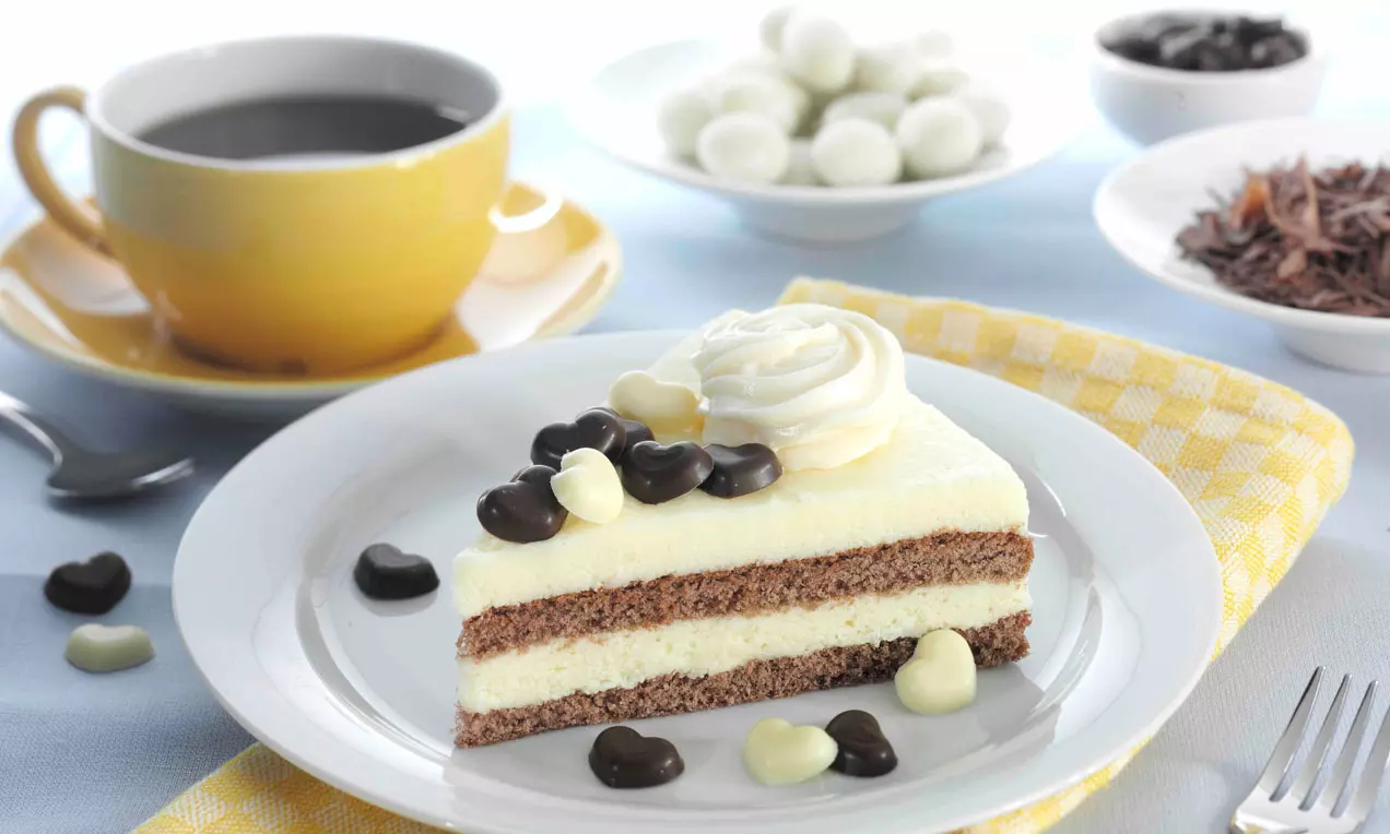 Ein dreieckiges Stück Buttercremetorte mit hellgelber Creme und braunen Böden, dekoriert mit Sahnetupfen und dunklen sowie hellen Schokoladenherzen auf weißem Teller
