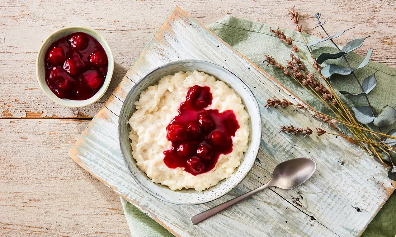 Cremiger weißer Milchreis mit glänzend roter Kirschgrütze in einer grauen Schale auf hellen Holzbrettern mit Grünzweigen und Löffel drapiert