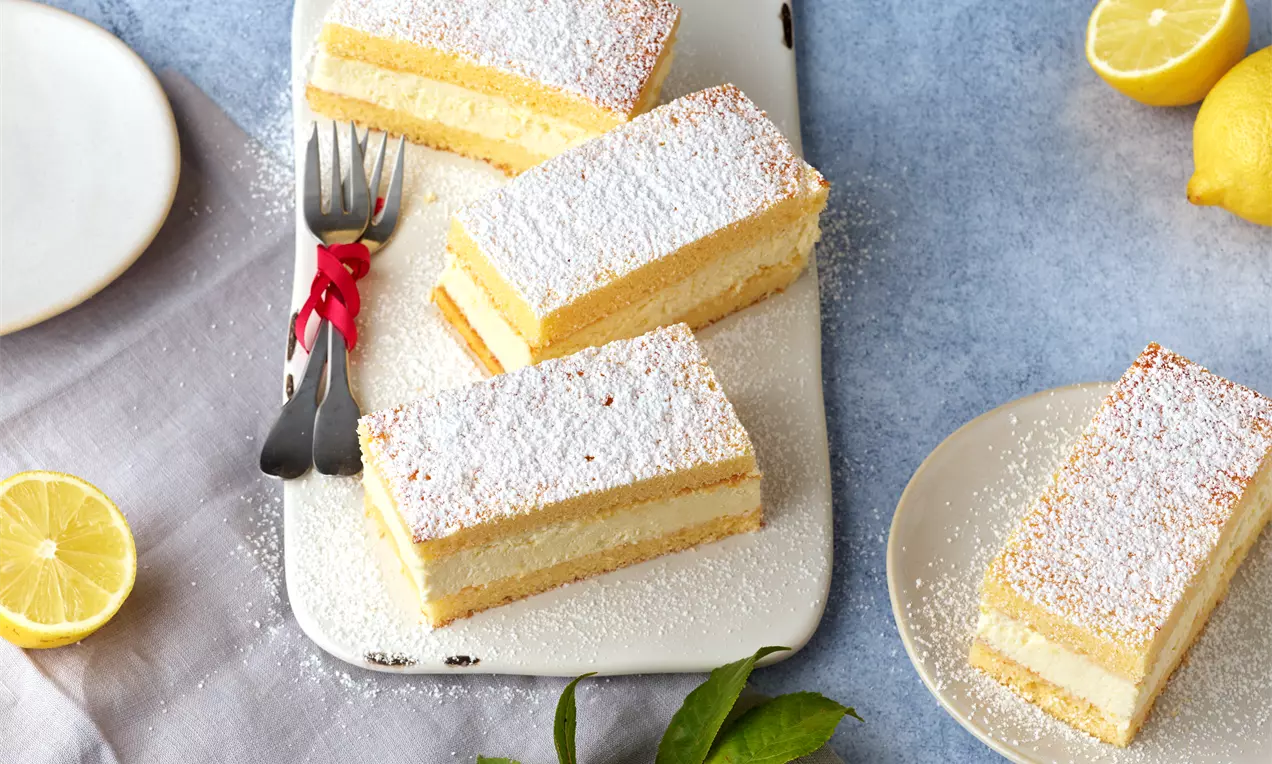 Rechteckige Zitronen-Mascarpone-Schnitten mit gelber Schichtfüllung und Puderzucker bestäubt auf heller Platte mit Zitronen dekoriert