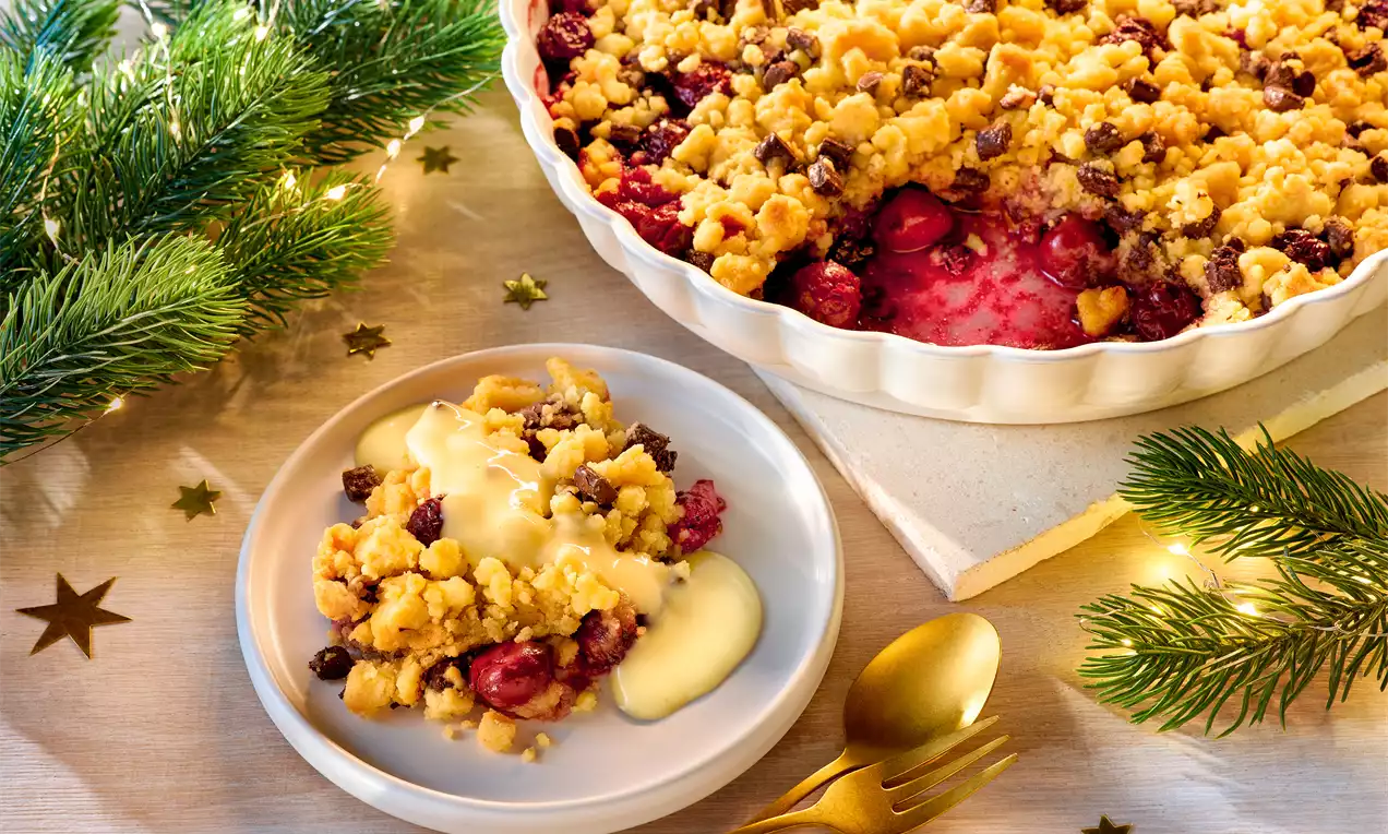 Weihnachts-Crumble mit goldbraunen Streuseln, roten Früchten und Vanillesoße, serviert auf weißem Teller mit goldfarbenem Besteck, dekoriert mit Tannenzweigen