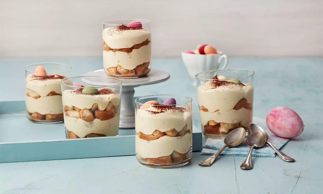 Eierlikör-Tiramisu im Glas mit geschichteter Creme, goldbraunen Löffelbiskuits, Schokostreuseln und bunten Zuckereiern auf einer blauen Fläche