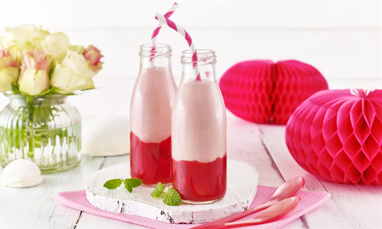 Zwei Glasflaschen mit rosafarbenem Joghurt-Shake auf roter Fruchtgrütze, serviert auf einem weißen Holzbrett mit rosa Dekoration