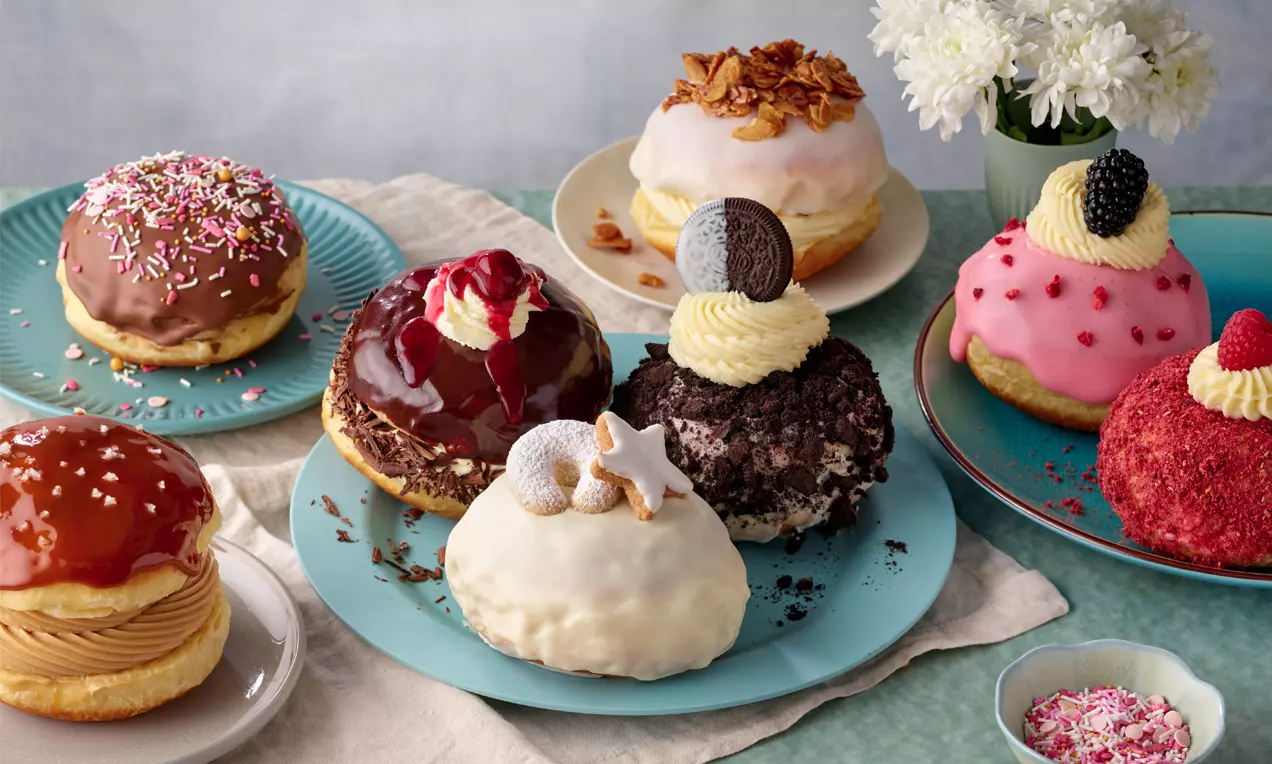 Verschiedenfarbige Krapfen mit glänzender Glasur, Cremetupfen und Dekoration, auf Tellern drapiert, umgeben von Blumen und kleinen Schüsseln