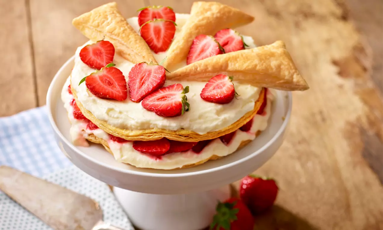 Auf einer weißen Tortenplatte geschichtete Blätterteigtorte mit cremeweißer Füllung und saftig roten Erdbeeren, dekoriert mit Teigdreiecken