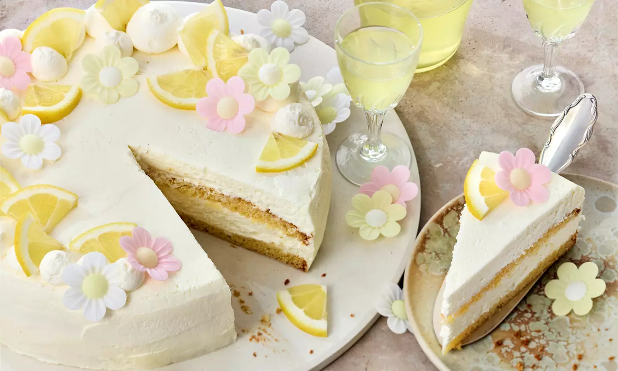 Limoncello-Torte mit hellgelber Creme, Zitronenscheiben und bunten Blüten auf hellem Teller, ein angeschnittenes Stück auf beigem Teller