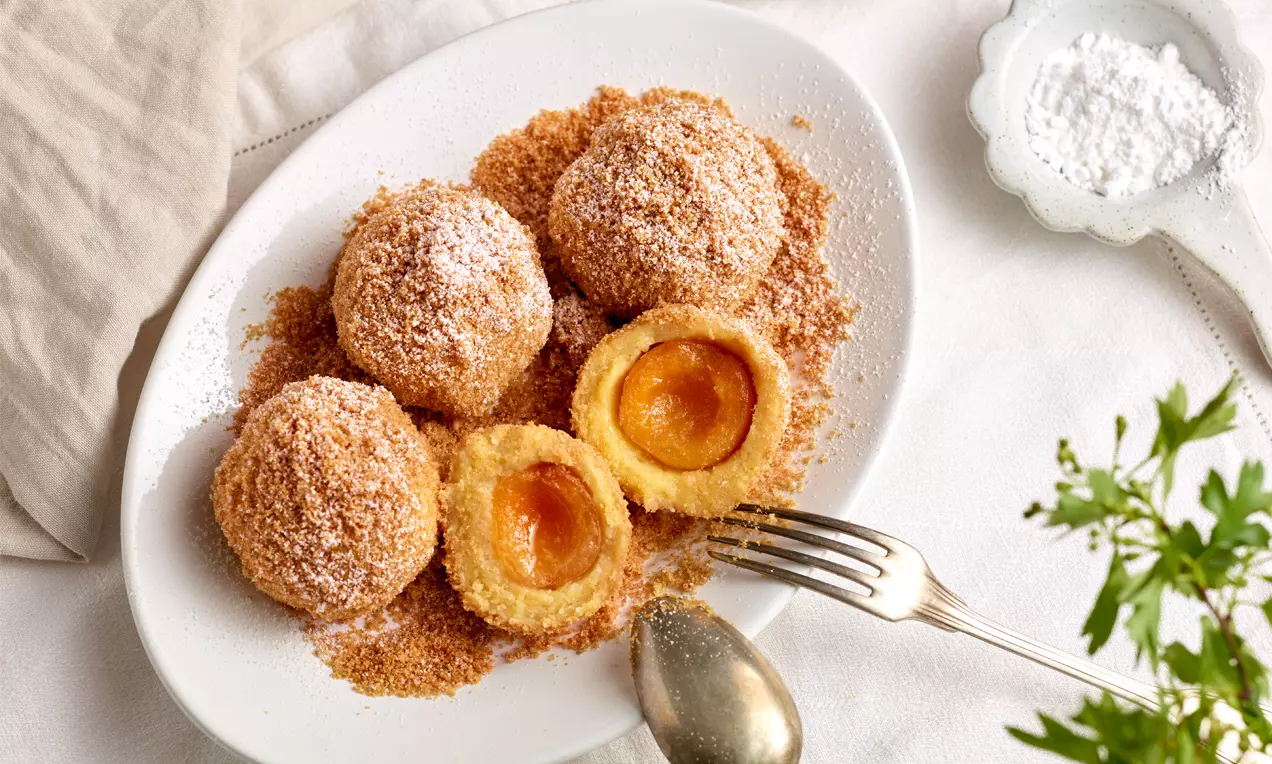 Goldbraune Marillenknödel in knusprigen Bröseln, ein Knödel halbiert mit saftiger orangefarbener Fruchtfüllung auf weißem Teller mit Besteck