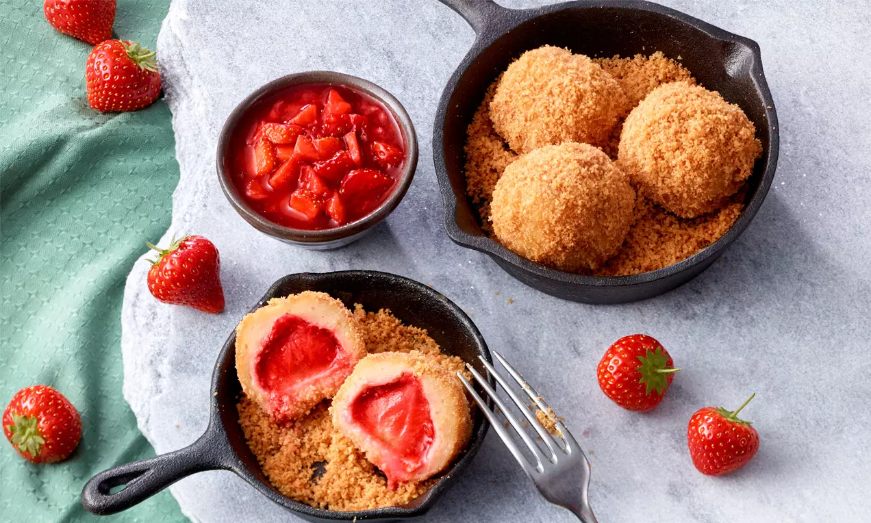 Erdbeerknödel mit goldbrauner Bröselkruste in schwarzen Pfannen, eine angeschnitten mit rotem Kern, umgeben von frischen Erdbeeren und Erdbeersauce