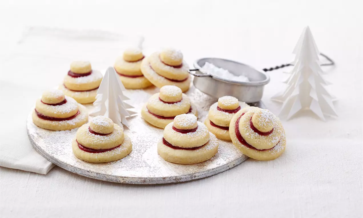 Terrassenplätzchen aus hellgoldenem Mürbeteig mit roter Füllung und Puderzucker bestäubt auf einem weißen Teller mit Papierdeko und Sieb