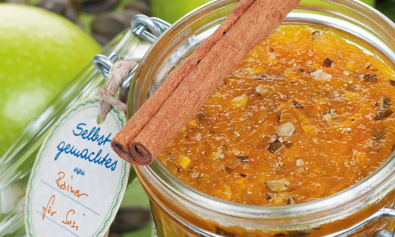 Leuchtend orange Apfel-Kürbismarmelade mit Stückchen in einem offenen Schraubglas, dekoriert mit einer Zimtstange und grünem Apfel im Hintergrund