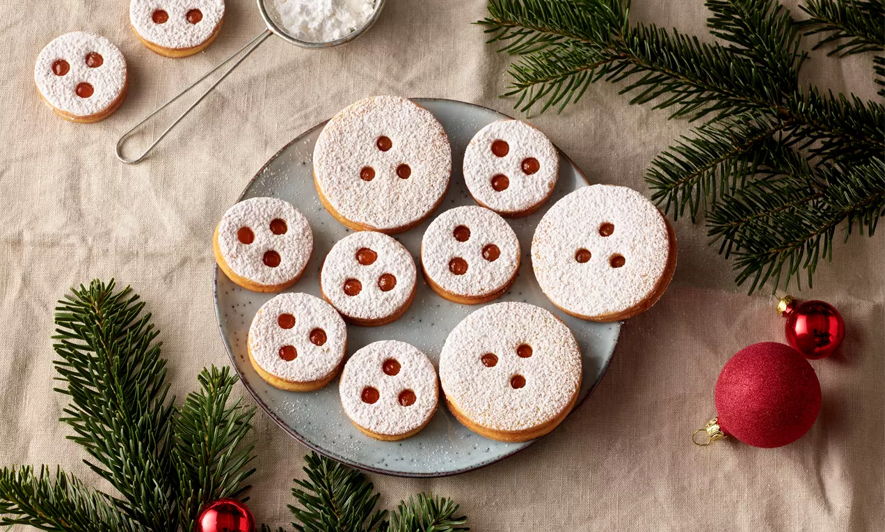 Runde Linzer Augen mit weißem Puderzucker und roter Marmeladenfüllung auf grauem Teller, dekoriert mit Tannenzweigen und Weihnachtskugeln