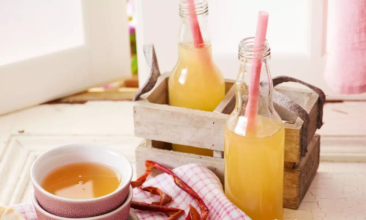 Naturtrüber Apfelsaft in klaren Glasflaschen mit rosa Strohhalmen, serviert in einer Holzkiste neben einer Tasse auf hellem Stoff
