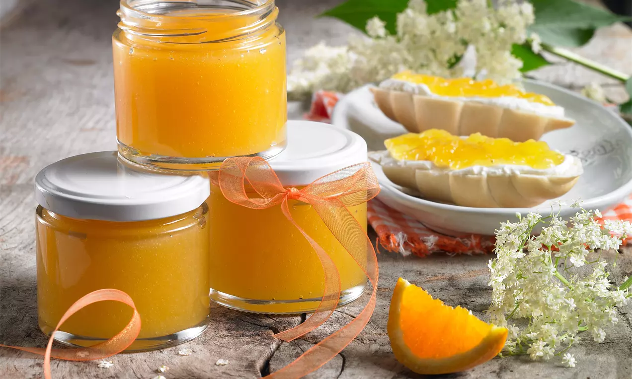 Drei Gläser mit leuchtend orangefarbenem, glattem Gelee auf rustikalem Holztisch, daneben weiße Blüten, Orangenschnitz und belegte Brötchen