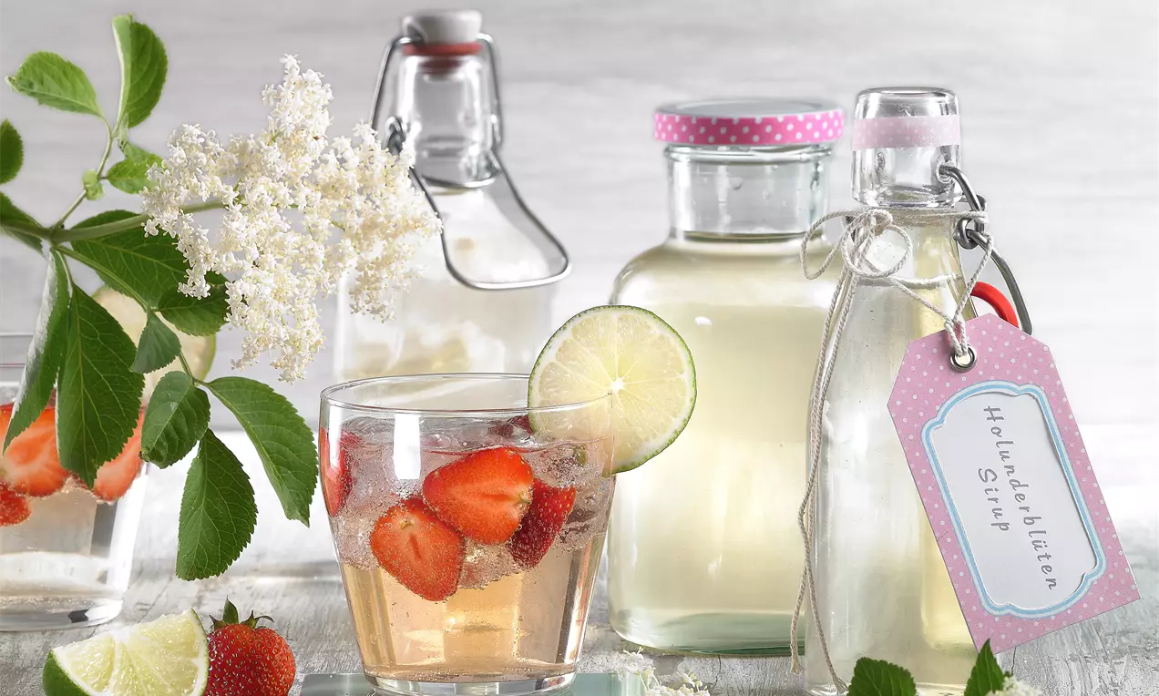 Helle Glasflaschen mit Holunderblütensirup und rosa Etiketten, davor ein Glas mit hellem Getränk, Erdbeeren, Limettenscheibe und Blütendeko