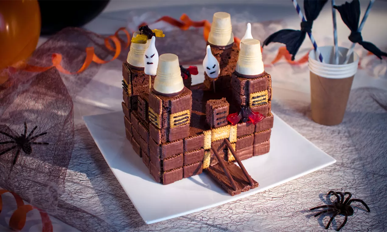 Schokoladenkeks-Schloss mit weißen Gespenstern aus Sahne, gelben Details und dekorativen Portalen auf weißer Platte, umgeben von Halloween-Deko