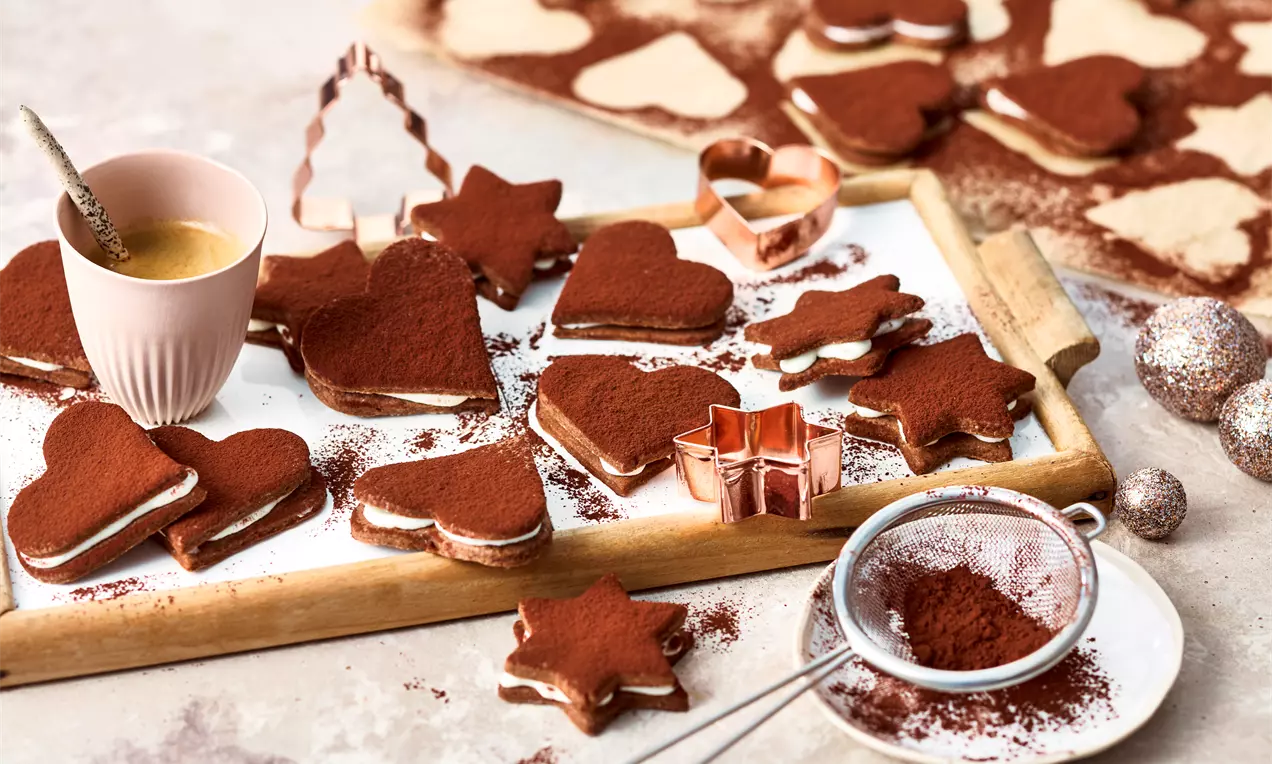 Herz- und sternförmige Tiramisu-Plätzchen mit kakaoüberzogener Oberfläche und heller Cremefüllung auf einem Holzbrett mit Deko und Sieb