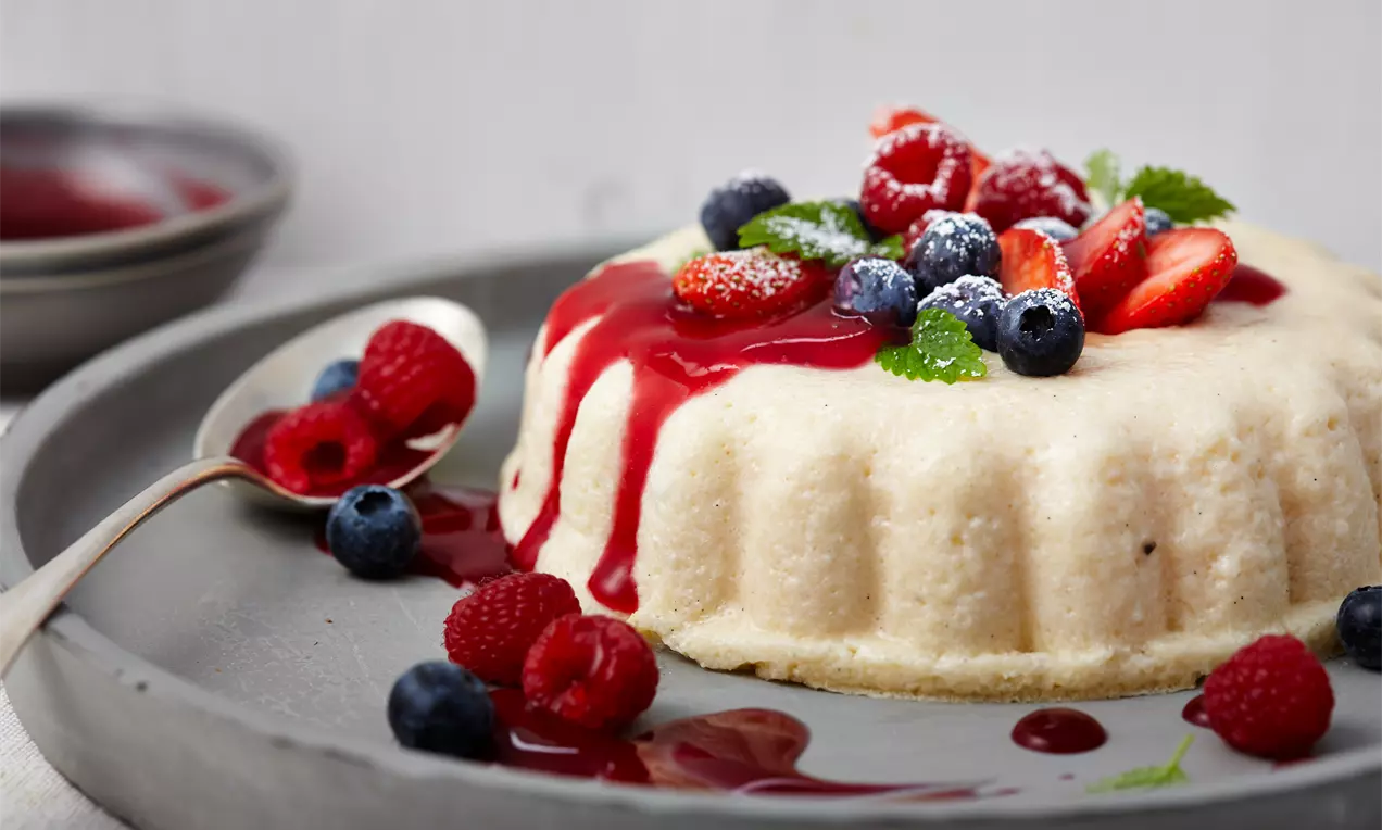 Heller Grießpudding mit glatter Textur, dekoriert mit roter Fruchtsoße, frischen Beeren und Minzblättern auf einem grauen Teller