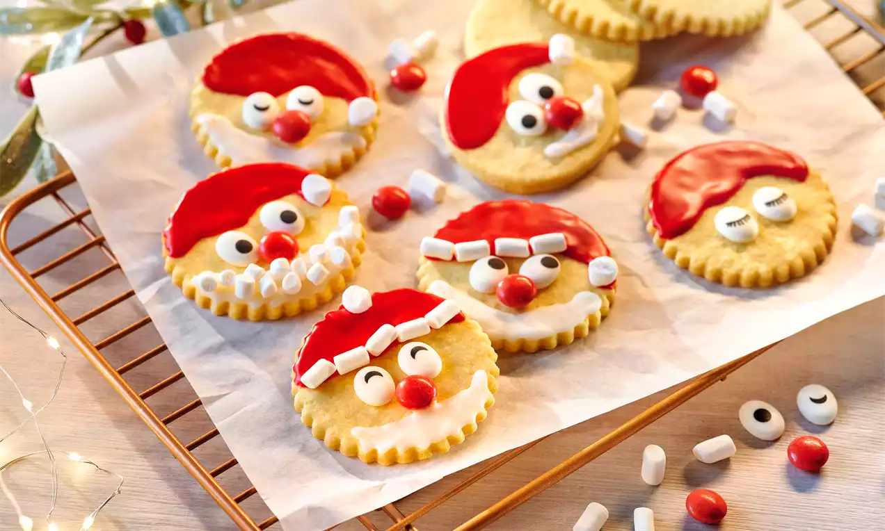 Runde, goldgelbe Butterplätzchen mit gezacktem Rand, dekoriert als Nikolausgesichter mit rotem Zuckerguss, Zuckeraugen und Marshmallows