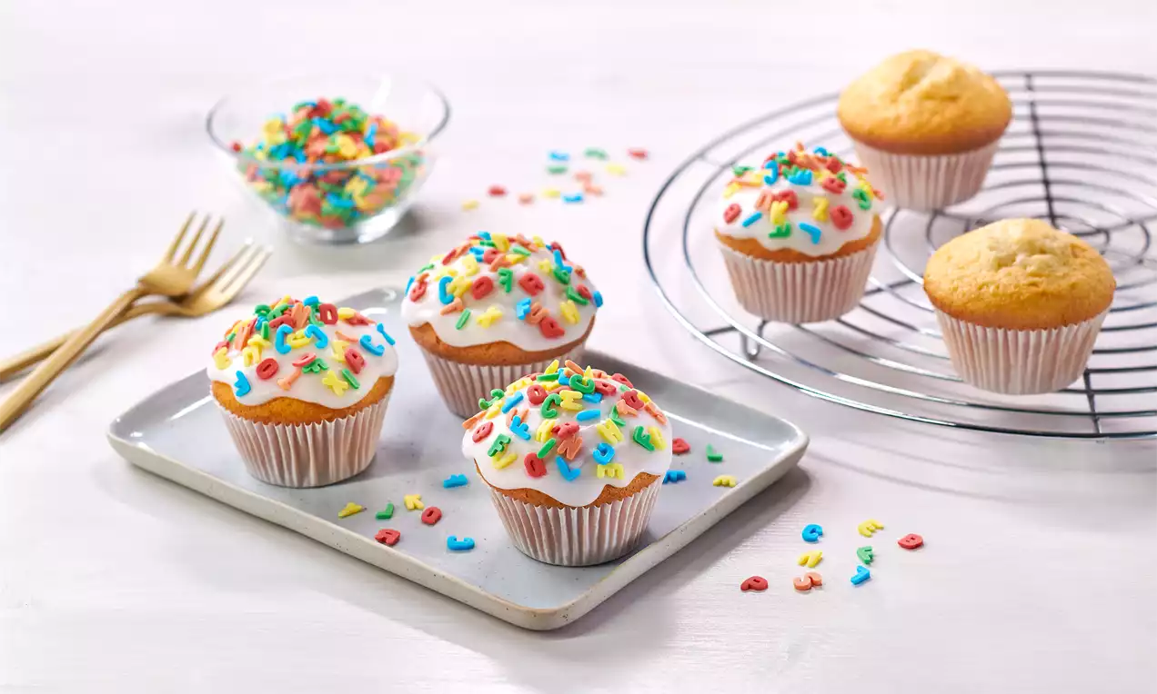 Goldgelbe Muffins mit heller Glasur und bunten Zuckerstreuseln dekoriert, auf einem Tablett und Kuchengitter arrangiert, daneben bunte Streusel
