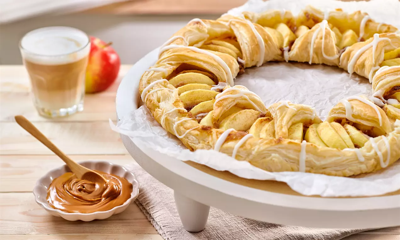 Runde Apfel-Tarte aus goldbraunem Blätterteig mit gefächerten Apfelscheiben und Zuckerguss auf weißem Teller, daneben Lotuscreme und Latte Macchiato