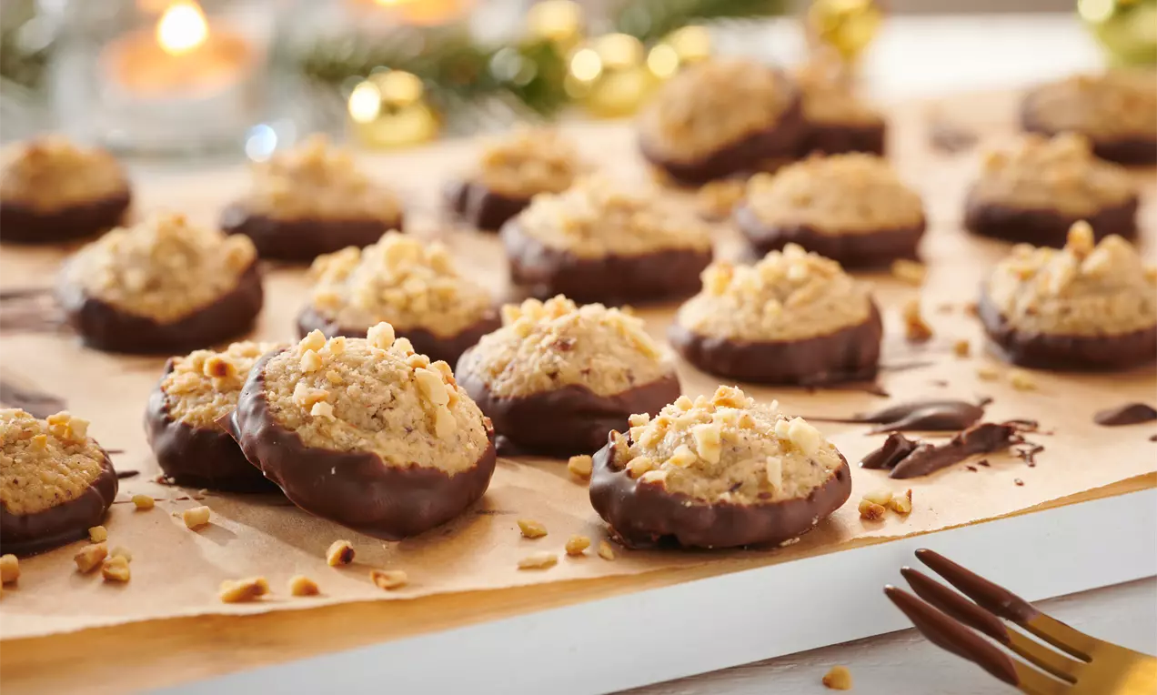 Goldbraune Marzipan-Makronen mit Haselnussstückchen, teils in dunkle Schokolade getaucht, auf Backpapier mit weihnachtlicher Dekoration im Hintergrund
