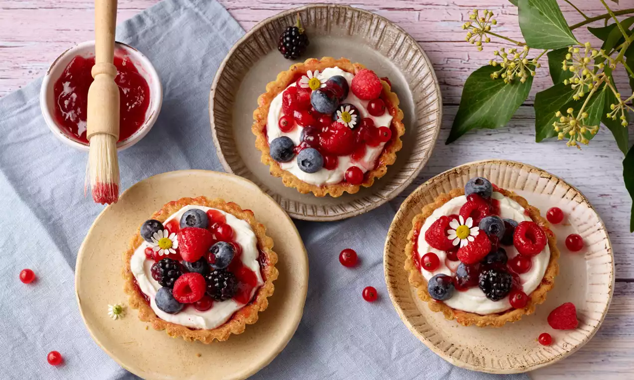 Kleine Tartelettes mit goldbraunem Mürbeteig, weißer Vanillecreme und frischen, glänzenden Beeren, dekoriert mit Kamillenblüten auf Tellern