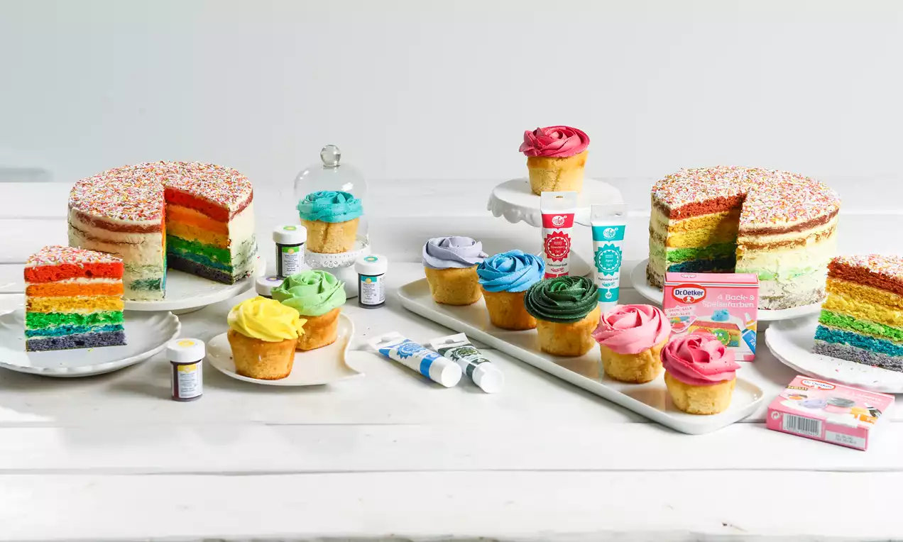 Regenbogen-Torte mit bunten Schichten und Zuckerstreuseln auf weißem Tisch, umgeben von farbiger Lebensmittelfarbe und Cupcakes