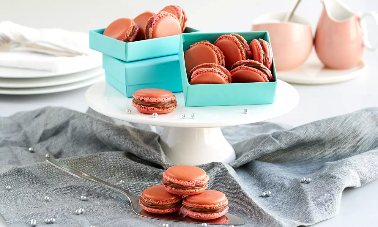 Rote Macarons mit glatter Oberfläche und zarter Nuss-Nougat-Füllung auf einer weißen Tortenplatte, umgeben von türkisfarbenen Boxen