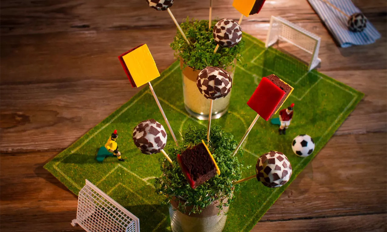 Fußball-Cakepops mit braun-weißer Musterung und farbigen Würfeln an Holzspießen dekorativ auf Rasen in Gläsern arrangiert