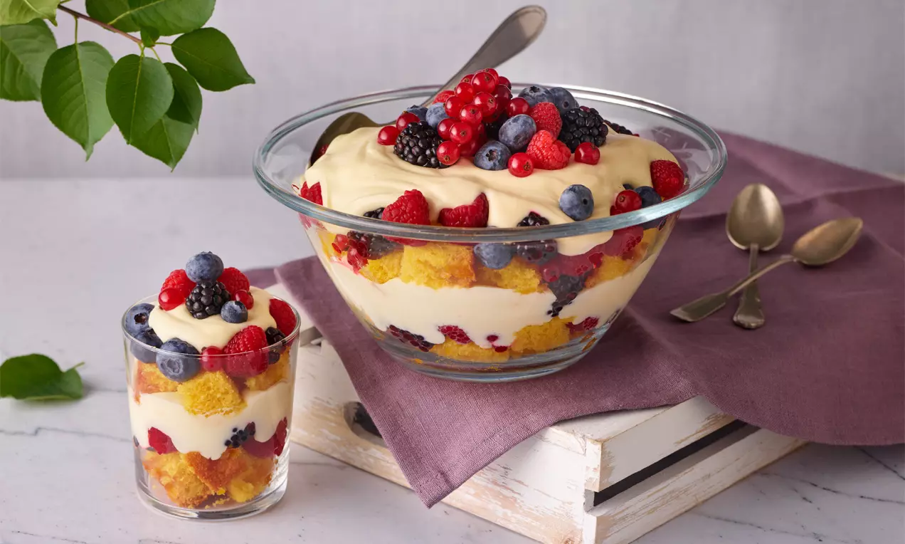 Schichtdessert aus heller Creme, saftigen Kuchenstücken und bunten Beeren in einer Glasschale, garniert mit Beeren und Löffeln daneben