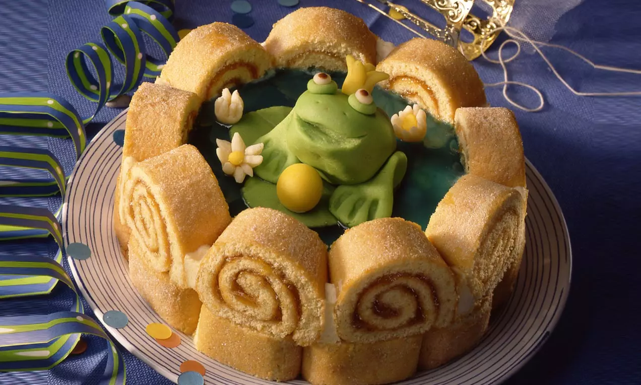 Eine Froschkönig-Torte aus goldenen Biskuitrollen, gefüllt mit grünem Marzipan-Frosch auf blauem Spiegel, dekoriert mit Blüten und einer goldenen Krone