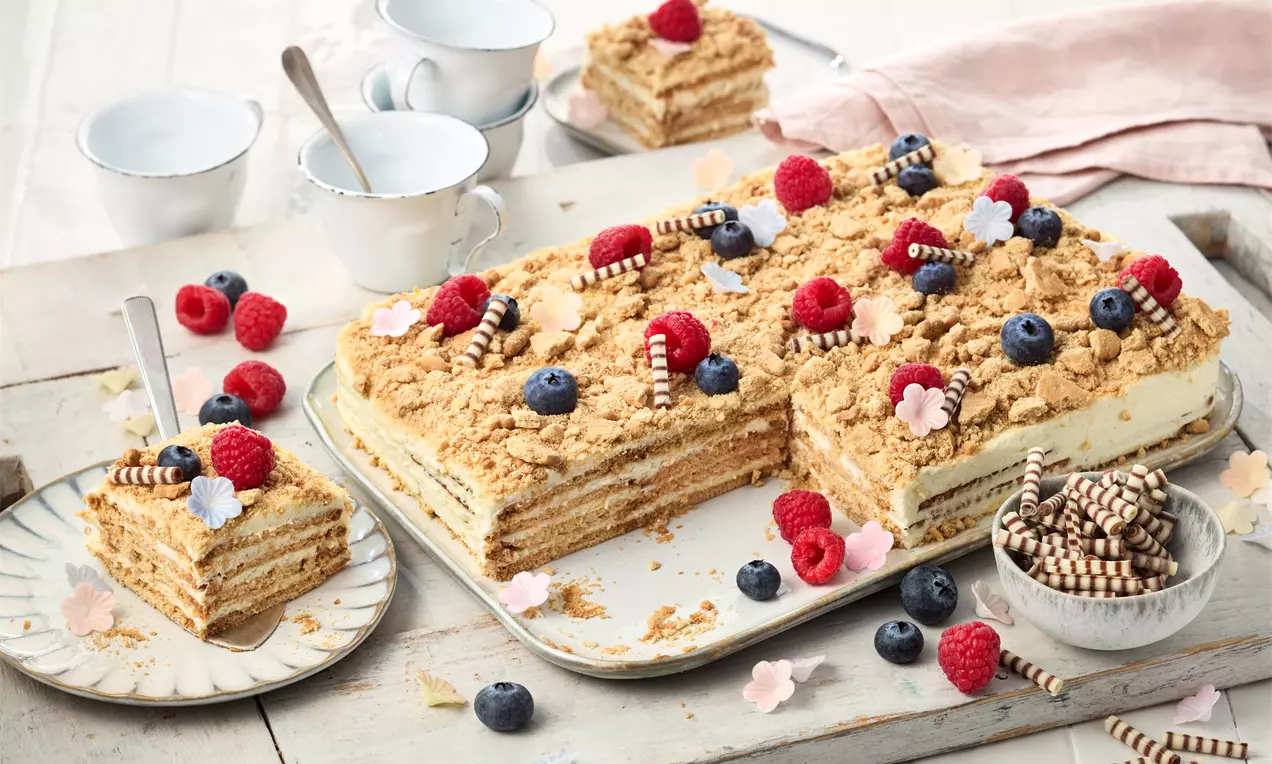 Rechteckiger Kuchen mit heller Creme, luftigen Schichten und knusprigem Streuseltopping, dekoriert mit Beeren, Schokoröllchen und Blüten