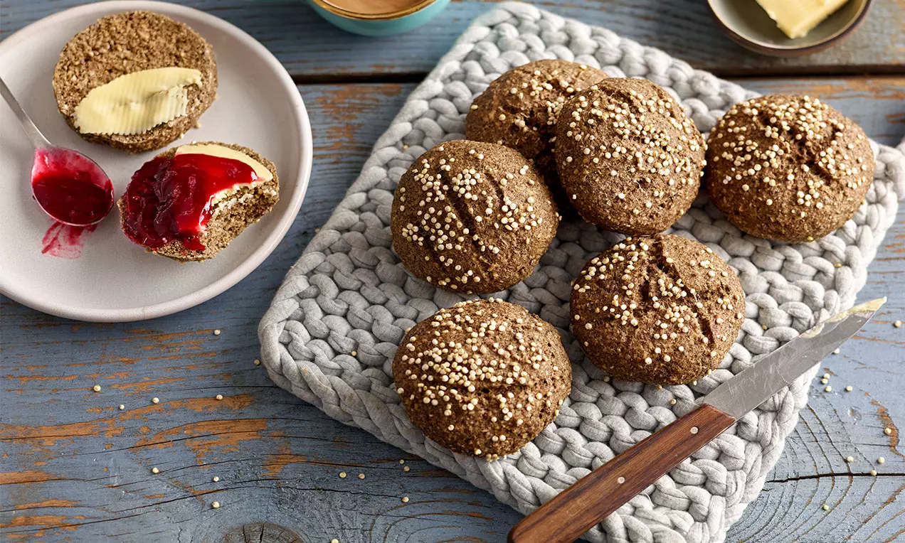 Runde, goldbraune Low-Carb-Brötchen mit knuspriger Kruste und weißen Körnern auf grauer Unterlage neben einem Teller mit belegten Hälften