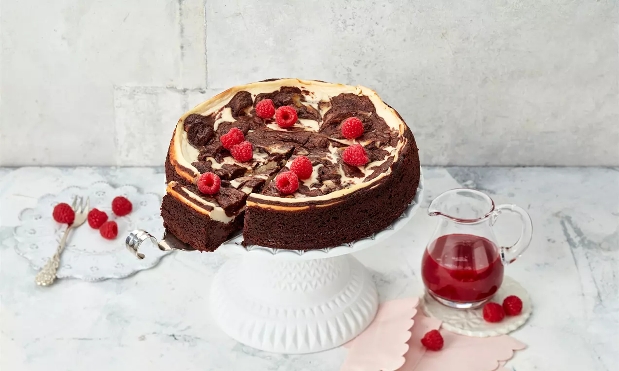 Brownie-Käsekuchen mit weißer Creme-Schicht und schokoladigem Marmor, garniert mit Himbeeren auf einer weißen Tortenplatte mit roter Sauce
