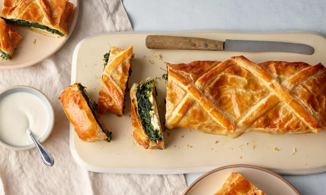 Goldbrauner Strudel mit filigranem Muster, gefüllt mit grüner Spinat-Schafskäse-Mischung, angerichtet auf einem rechteckigen Teller mit Messer