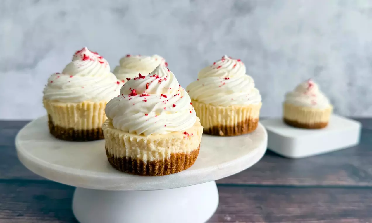 Kleine Cheesecakes mit goldbraunem Keksboden und weißem Schlagschaum, garniert mit roten Krümeln, auf einer weißen Tortenplatte drapiert, AI-labeled image