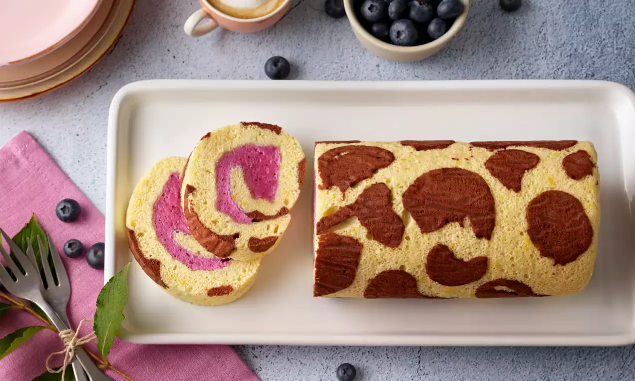 Heidelbeerroulade mit gelbem Teig und braunem Muster, gefüllt mit lilafarbener Creme, angeschnitten auf weißer Platte mit Deko aus Heidelbeeren