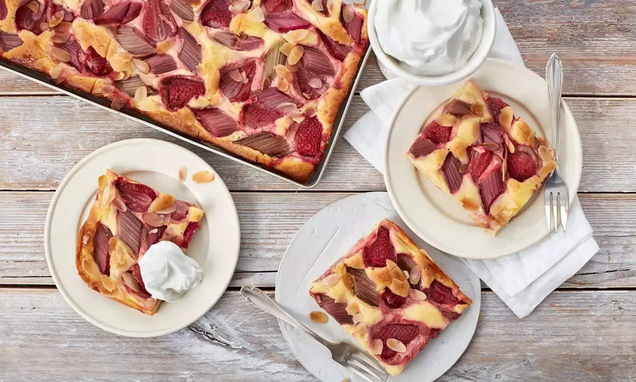 Erdbeer-Rhabarber-Kuchen mit goldbraunem Boden und fruchtig rotem Belag, garniert mit Mandelscheiben, serviert auf weißen Tellern mit Sahne
