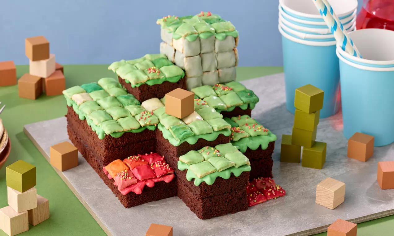 Minecraft-Kuchen in Würfelform mit braunem Schokoboden und bunter Pixelglasur, dekoriert mit Streuseln, Holzklötzen und Bechern im Hintergrund