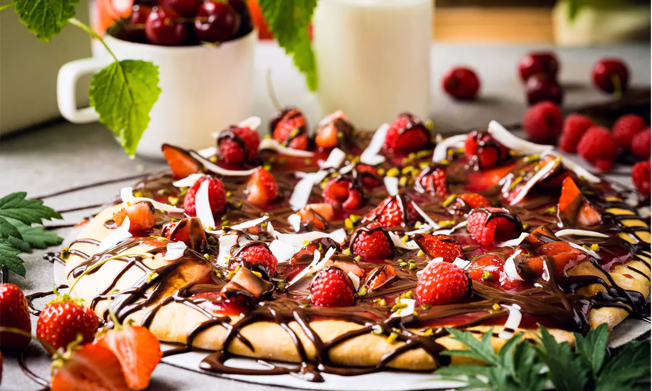 Runde, goldbraune Pizza mit glänzender dunkler Schokoladensauce, frischen roten Beeren, Kokosraspeln und grünem Dekor auf grauem Untergrund