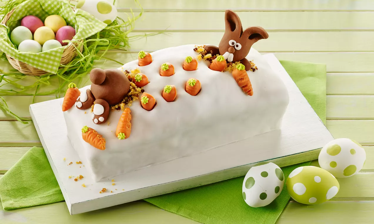 Weiß glasierter Möhren-Marmorkuchen mit bunten Fondantmöhren und braunen Fondanthäschen auf grünem Stoff und heller Holzplatte