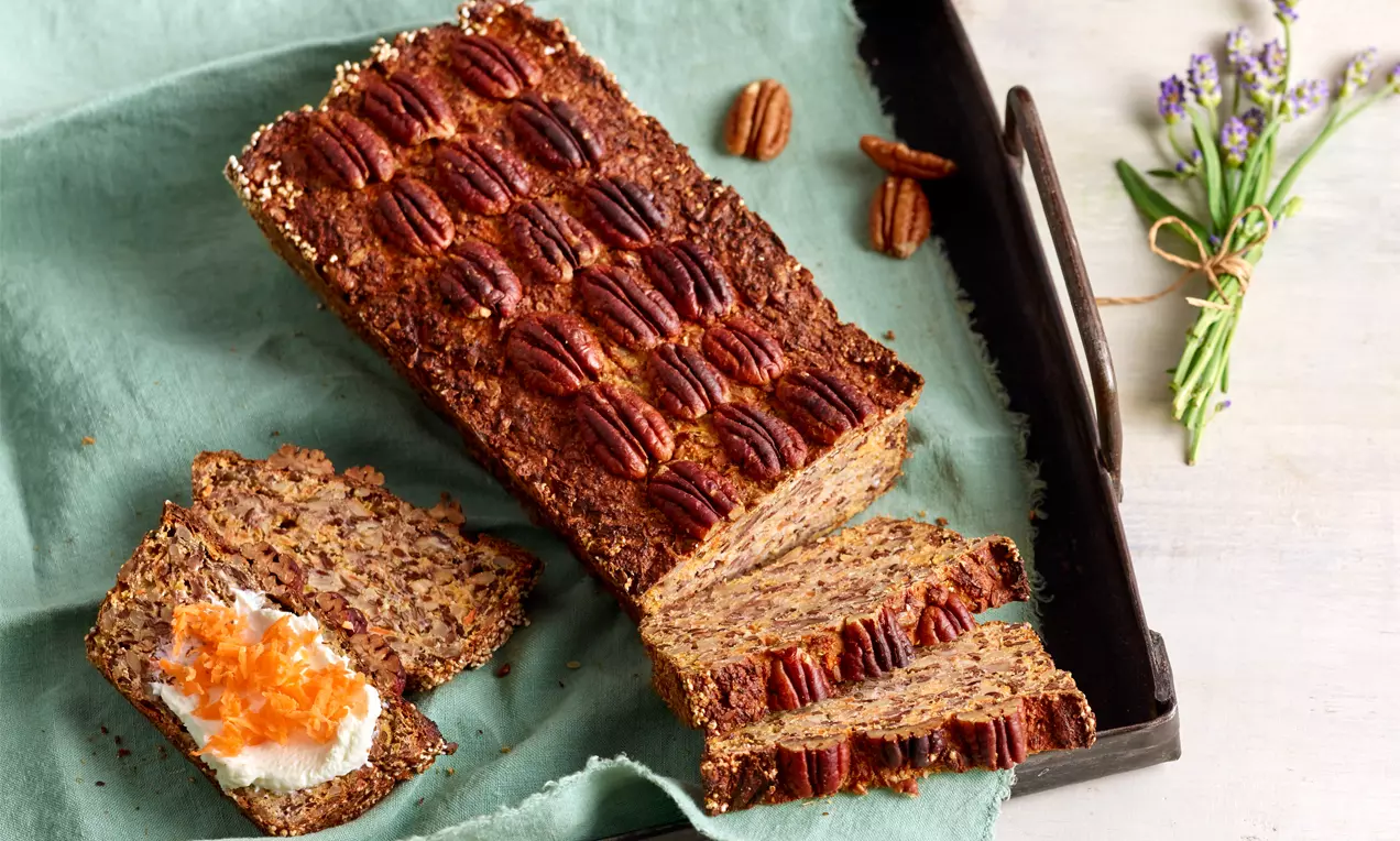 Rechteckiges, goldbraunes Eiweißbrot mit Pecannüssen belegt, eine Scheibe mit Frischkäse und Karottenraspeln auf grünem Tuch drapiert