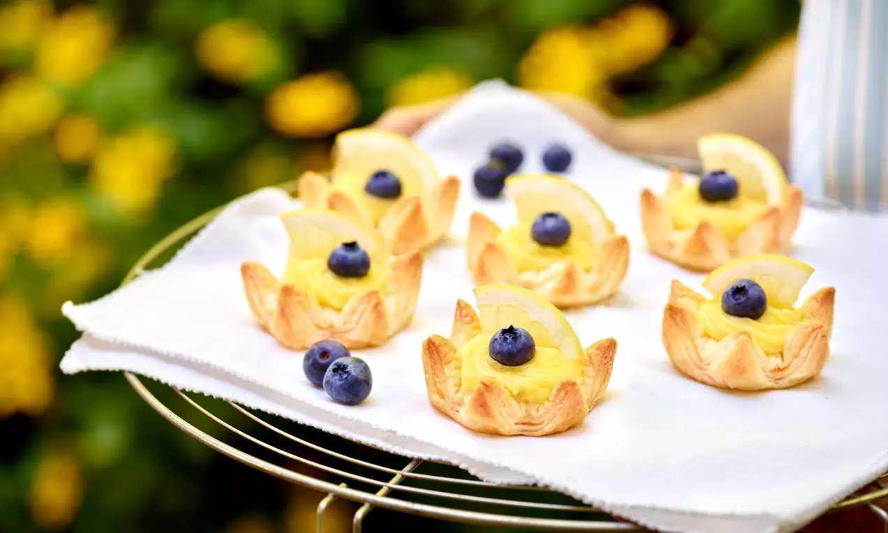 Goldbraune Blätterteig-Krönchen mit gelber Creme, dekoriert mit Zitronenscheiben und blauen Heidelbeeren auf weißem Tuch