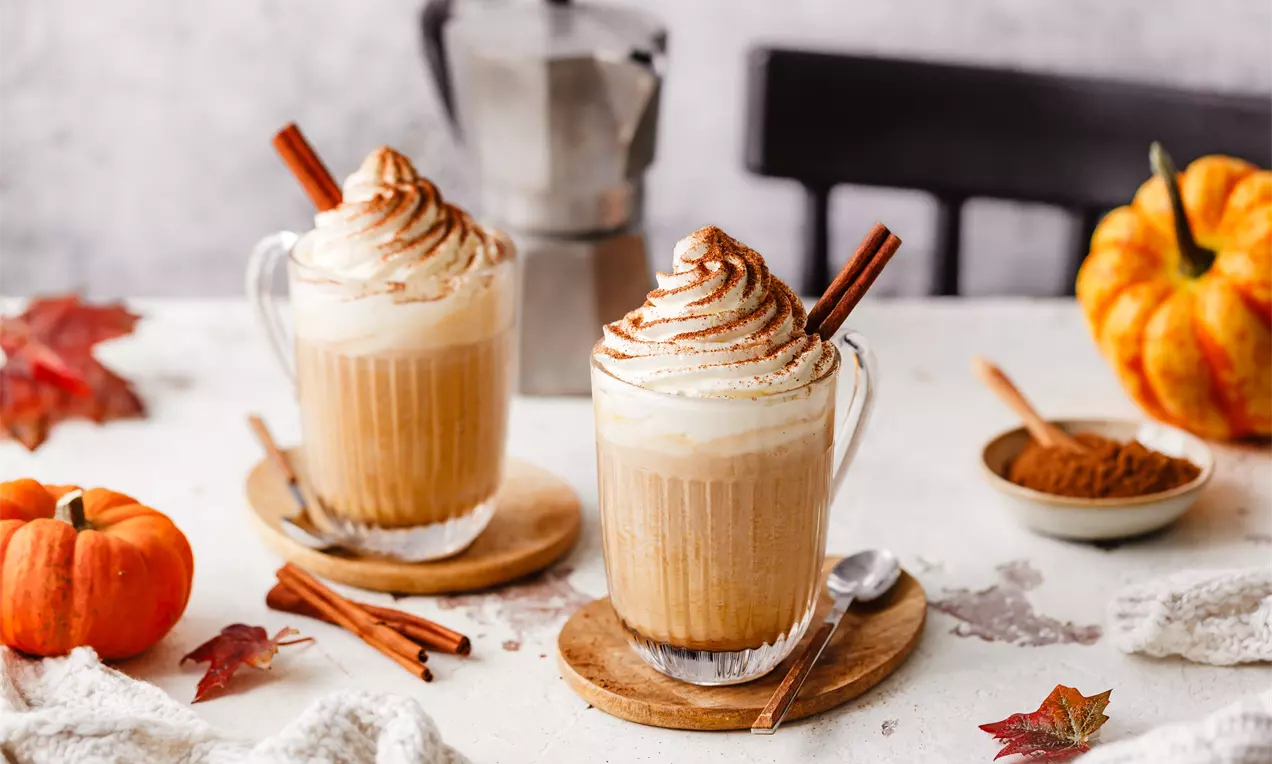 Zwei Glasbecher mit cremigem Pumpkin Spice Latte, getoppt mit Sahnehaube und Zimtstange auf Holzuntersetzern vor herbstlichem Hintergrund
