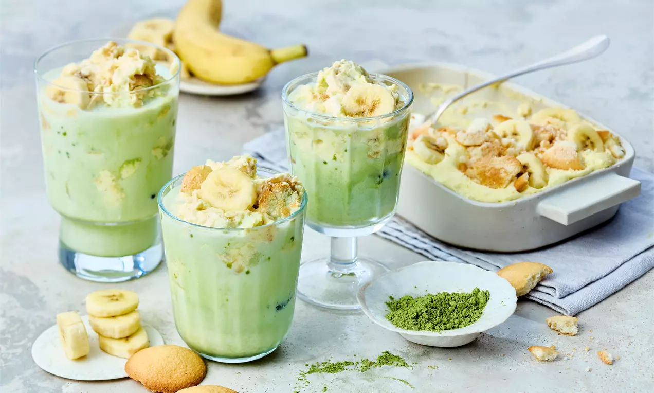Matcha-Bananen-Pudding in Gläsern mit grüner Creme, Bananenscheiben und Keksstücken, daneben Matcha-Pulver, Bananen und Auflaufform