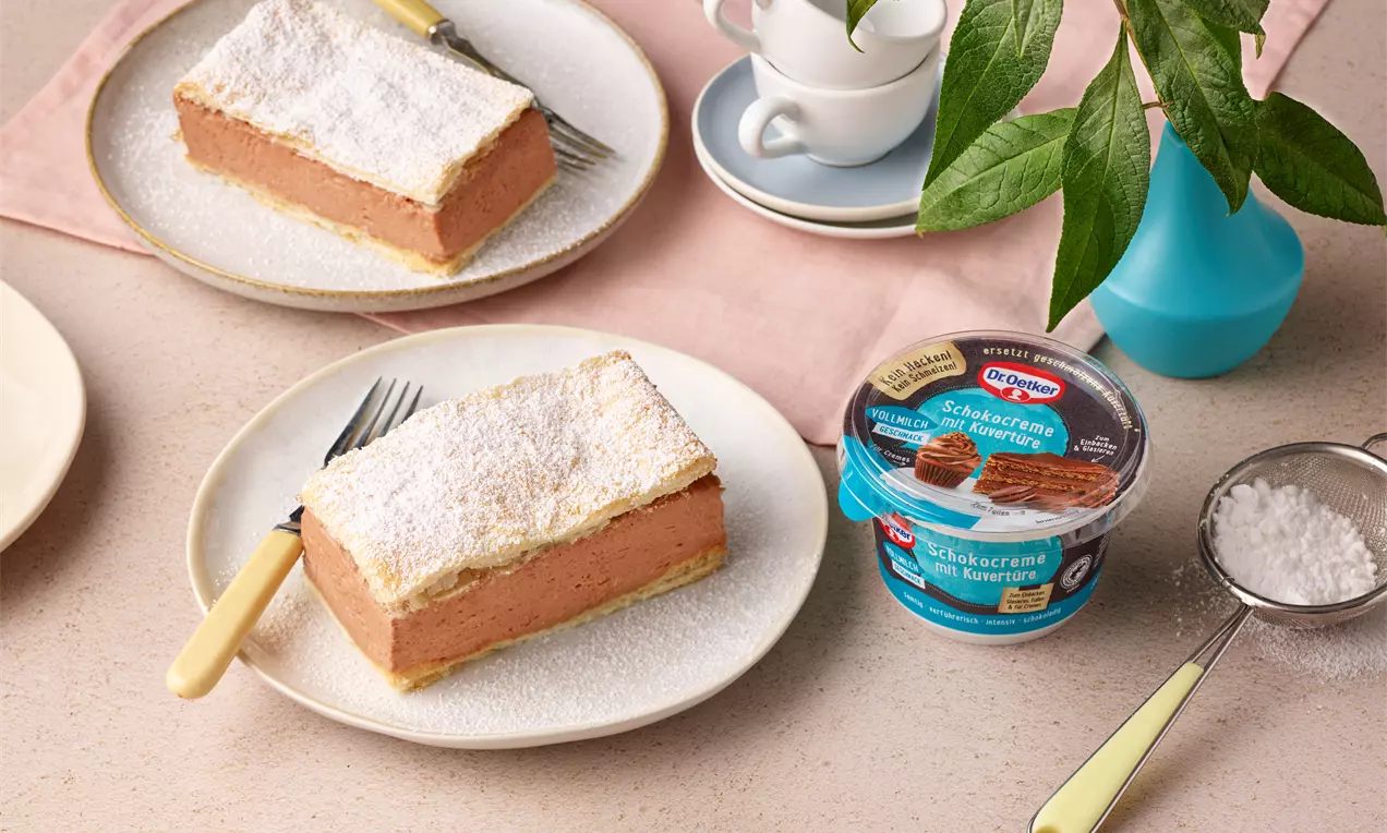 Rechteckige Schoko-Cremeschnitte mit heller, pudrig bestreuter Teigdecke auf einem weißen Teller, dekoriert mit gelbem Besteck