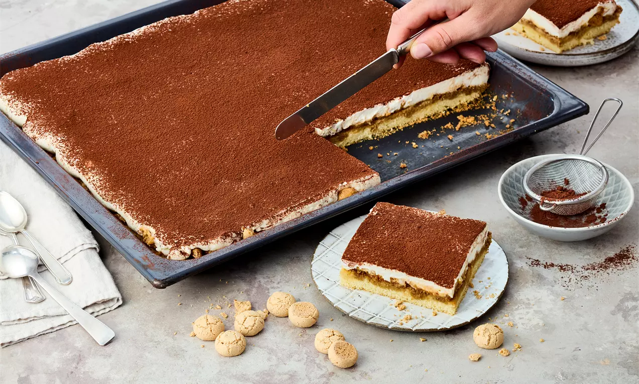 Tiramisu vom Blech mit brauner Kakao-Decke, heller Creme und luftiger Teigschicht, angerichtet auf Tellern mit Amarettini und Sieb