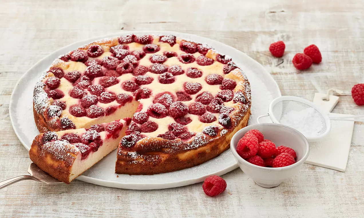 Himbeer-Käsekuchen ohne Boden goldbraun mit cremiger Oberfläche, dicht belegt mit frischen Himbeeren, bestäubt mit Puderzucker, auf weißem Teller