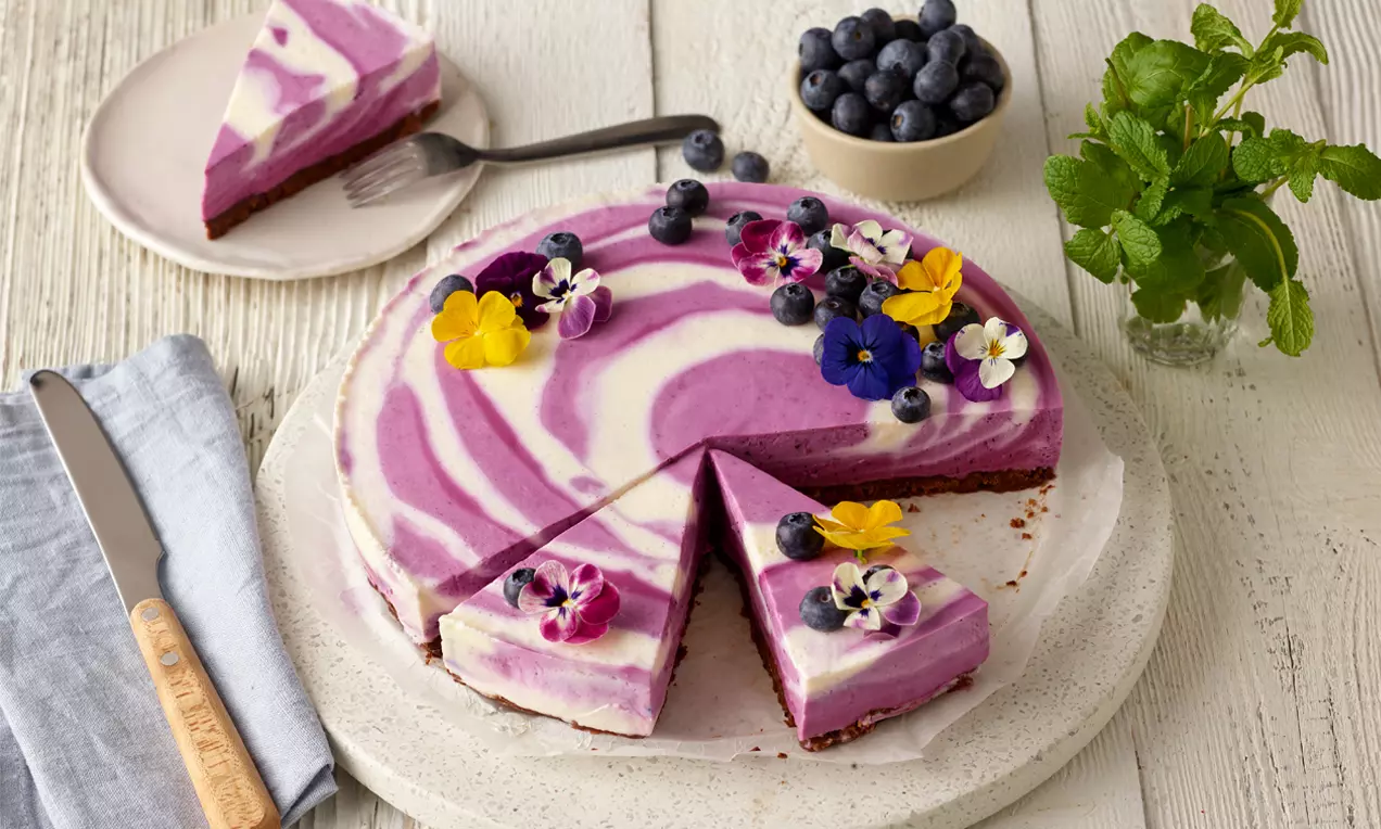 Cremiger No-bake-Cheesecake mit lila-weißer Swirl-Marmorierung, dekoriert mit Heidelbeeren und essbaren Blüten auf weißem Teller