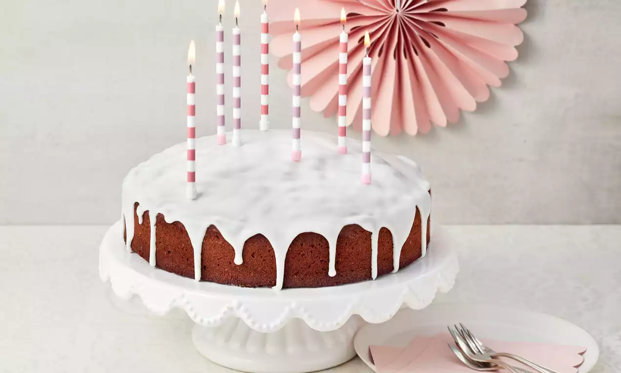 Runder Zitronenkuchen mit weißem Zuckerguss und rosa-weiß gestreiften Kerzen auf einer weißen Tortenplatte, daneben rosa Servietten mit Besteck